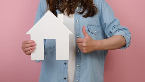 close up of unrecognizable young woman pointing on white paper house model and makes like gesture, real estate home approval, isolated on pink studio background. concept of social service about home