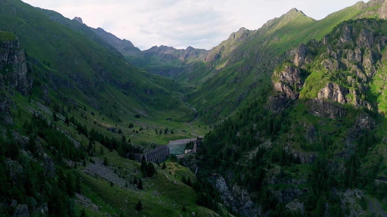 Aerial flight through the steep, forested gorge of Val di Scalve during sunset, approaching the historic Gleno Disaster Dam ruins