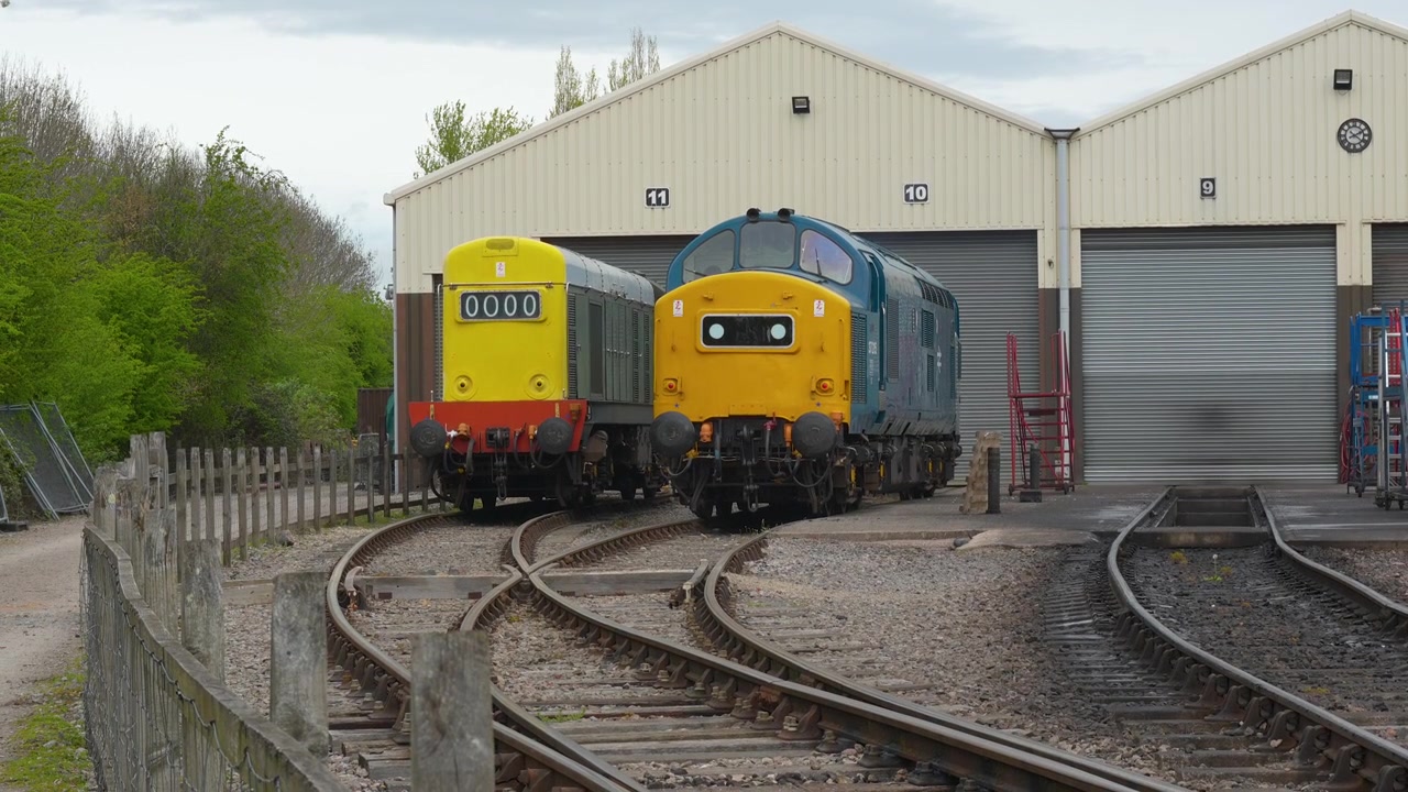 To klassiske britiske diesellokomotiver, herunder et Class 37, står stille på spor uden for et vedligeholdelsesdepot. Velegnet til dokumentarer om historisk transport og jernbanearv