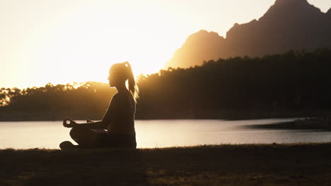 silhouette of woman meditating doing yoga by beautiful lake and mountains at sunset