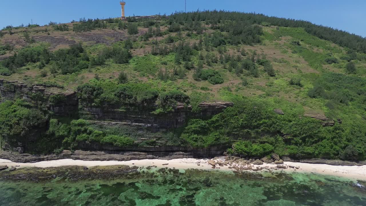 Drone zoom in showing sharp rock layers, green foliage and coral reef on clear beach coast