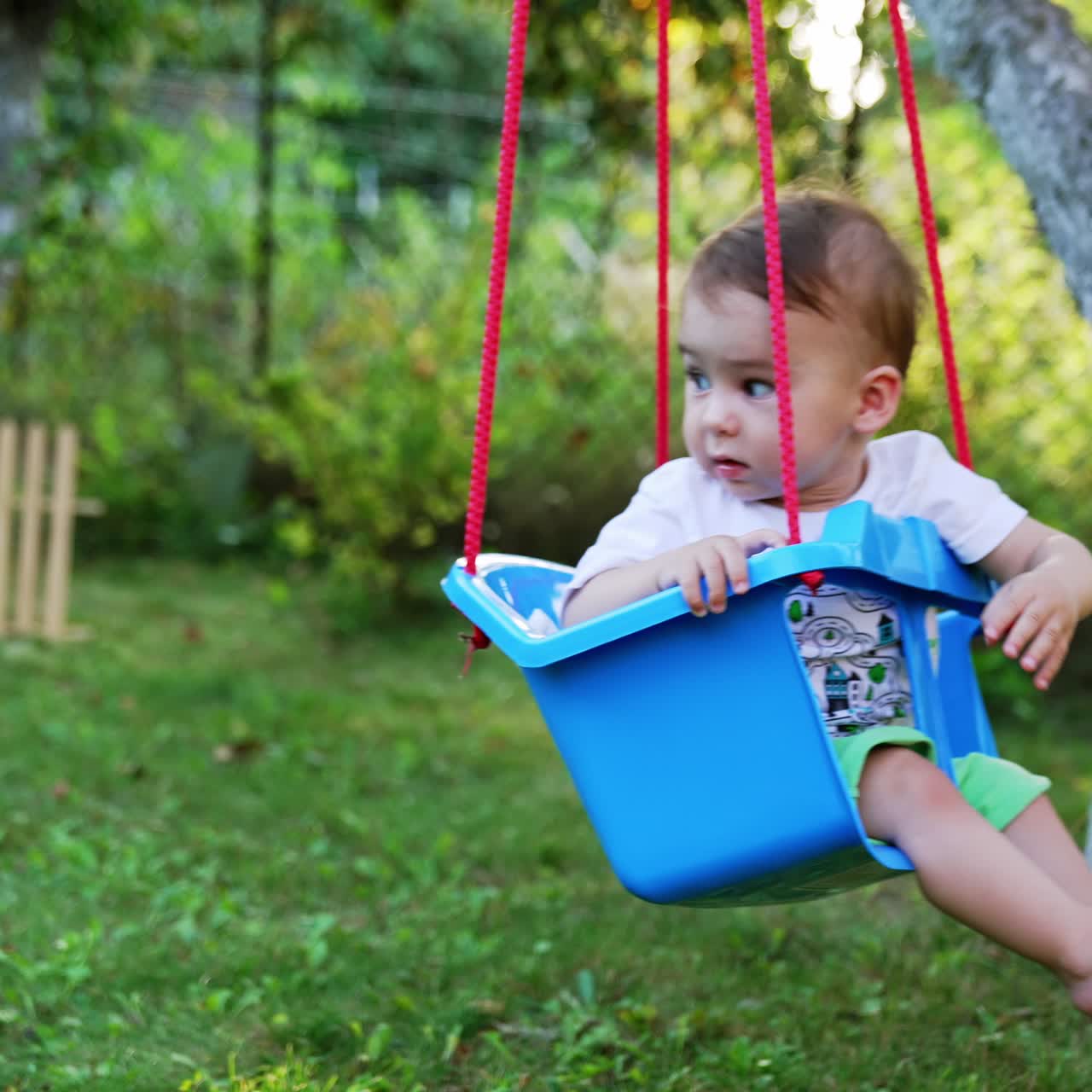 Lovely barefoot Caucasian boy swinging in a blue swing hanging from a tree. Toddler waving hand happily swaying. Garden background