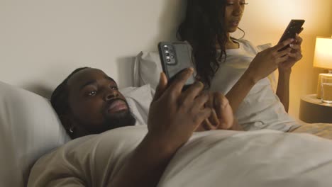 couple with relationship problems at home at night lying in bed both looking at mobile phones 1