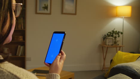 rear view of young woman spending evening at home sitting on sofa looking at blue screen mobile phone 1