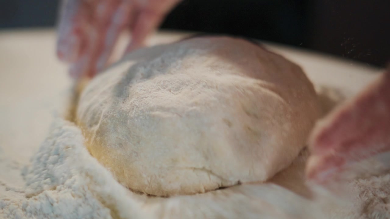 Hand drops dough into flour. Hands knead dough gently. Process of making bread or pastry shown. Can be used for cooking, baking, food blogs, recipes, or culinary education content
