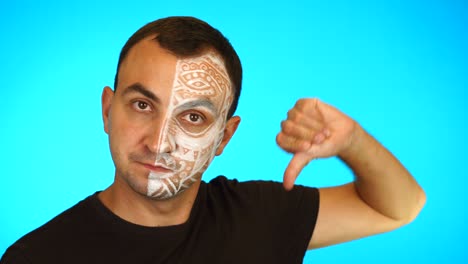 man with painted face showing thumb down and looking at camera against blue background