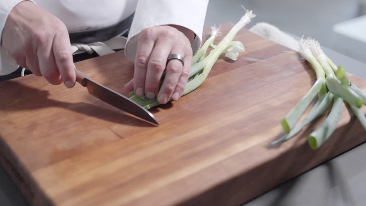 A static shot captures a chef finely chopping green onions on a cutting board, filmed at 120 fps to emphasize precise knife skills and culinary preparation.