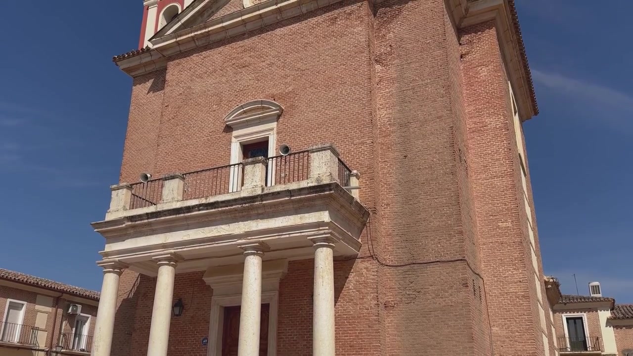 Movimento di telecamera verso l'alto che rivela la Cappella Reale di San Isidro nel Cortijo de San Isidro, mostrando il suo portico in pietra calcarea, il balcone, il frontone triangolare, i campanili gemelli e la torretta centrale rotonda.