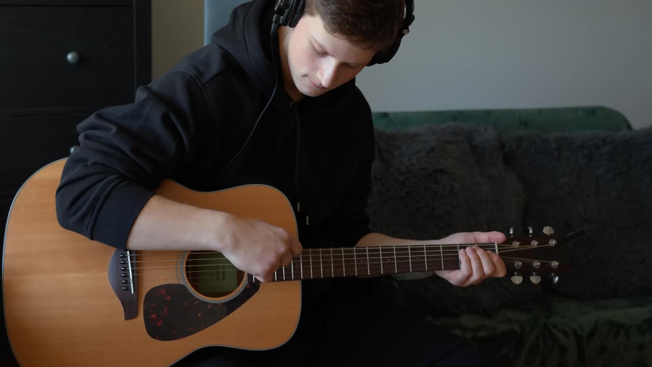 Fixed shot of a man wearing headphones and playing an acoustic guitar in a home setting, focused on strumming the strings while sitting on a couch