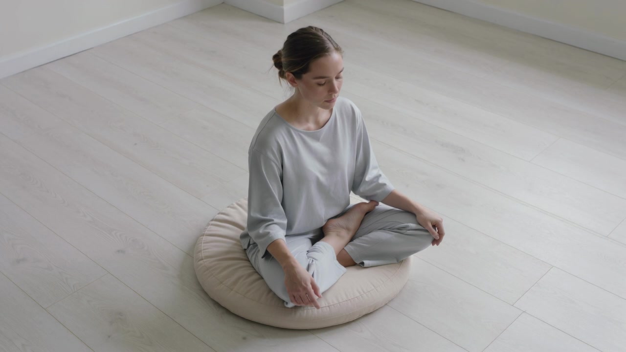 Donna in meditazione, in abbigliamento da casa grigio chiaro, seduta su un cuscino rotondo beige su un pavimento in legno, per un momento di calma.