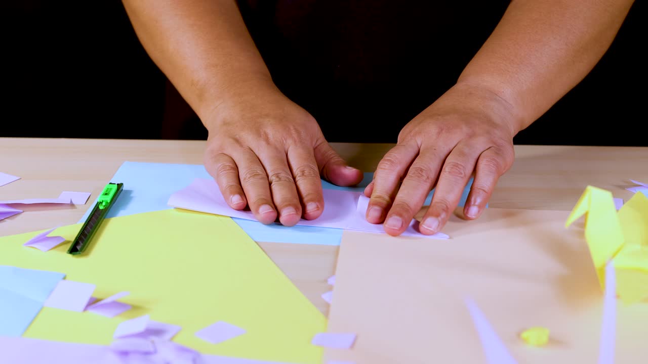 Person folds colored origami paper on tabletop under bright lighting, overhead camera, creative atmosphere