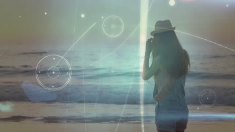 Woman in hat photographing ocean waves with digital network animation overlay