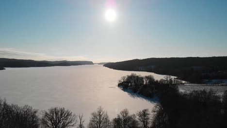 Frozen-Lake-Surrounded-By-Forest-Against-Bright-Sun-In-Blue-Sky