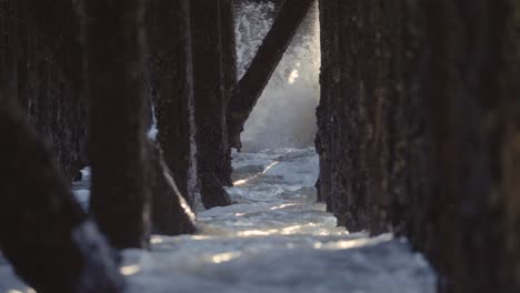 waves crashing under a pier, old wooden pillings with sunbeams coming through