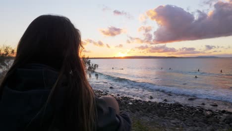 mujer mirando a los surfistas en la playa durante la puesta de sol en el parque nacional de noosa en australia