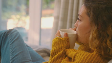 young woman in cosy warm jumper sitting on sofa at home looking out of window with hot drink