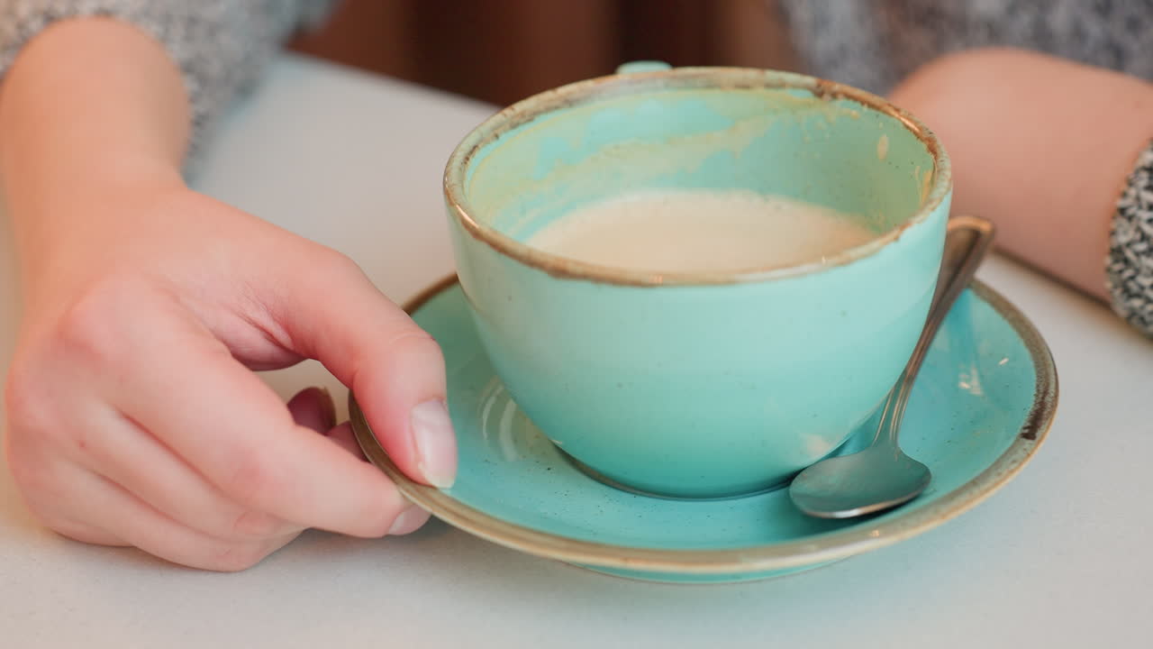 Fair lady gently moves hot tea cup sideways with fingertips on white table while enjoying cozy moment indoors with vintage ceramic saucer and spoon, conveying warmth and calm in relaxed atmosphere