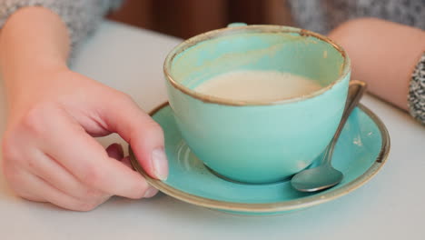 fair lady gently moves hot tea cup sideways with fingertips on white table while enjoying cozy moment indoors with vintage ceramic saucer and spoon, conveying warmth and calm in relaxed atmosphere