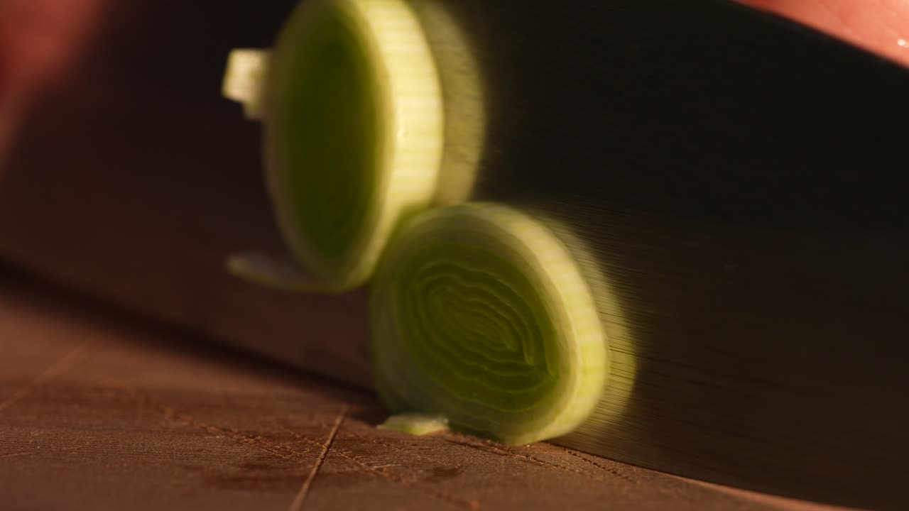 Close-up footage of a fresh leek being sliced on a wooden cutting board. The knife reveals crisp green layers, showing detailed food preparation and clean culinary technique