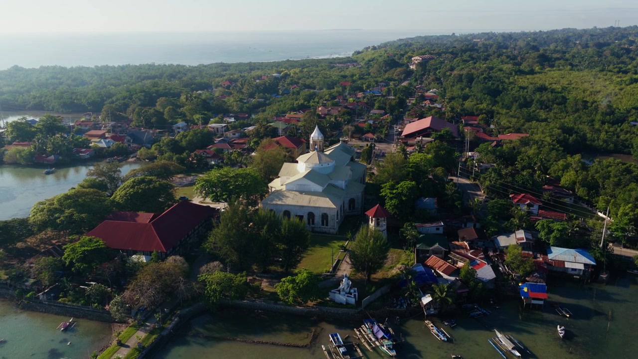 Gereja Dauis dikelilingi rumah-rumah tropis dan garis pantai, drone mengorbit dari tampilan belakang.