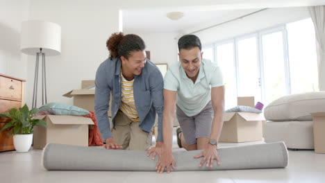 Happy-diverse-gay-male-couple-moving-into-new-home,-unrolling-rug-in-living-room,-slow-motion