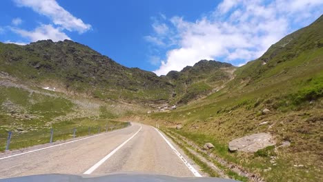 a car’s point of view on the transfagarasan mountain highway, surrounded by tall and green mountain peaks with patches of snow and a clear blue sky, transylvania, romania