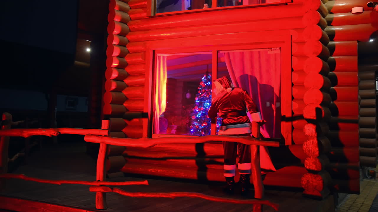 Santa Claus walking near the house. Father Frost at Christmas outdoors. Santa carrying presents at dark. Red lighting. Back view