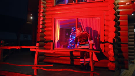 santa claus walking near the house. father frost at christmas outdoors. santa carrying presents at dark. red lighting. back view