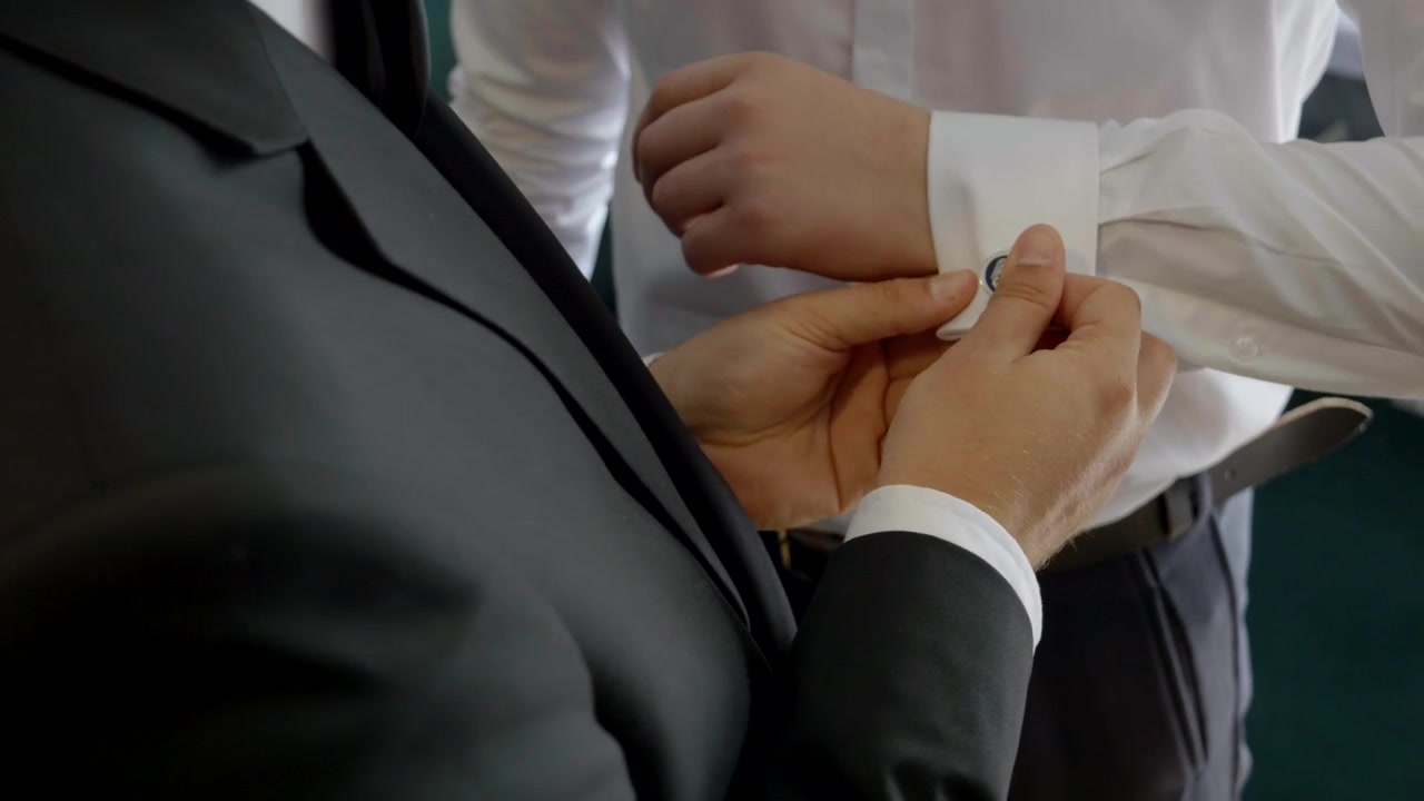 An elegantly dressed best man is adjusting the cufflinks on the groom's shirt cuffs during the wedding preparations