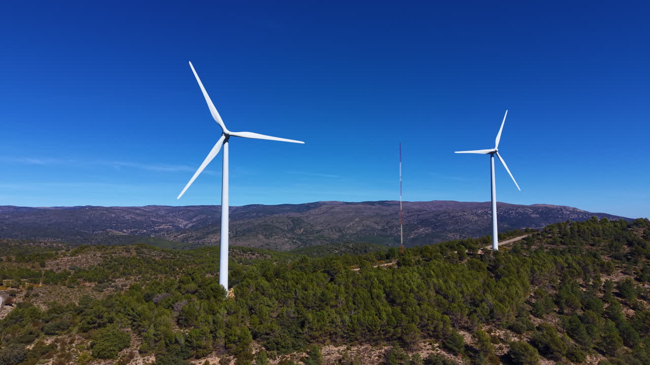 Due turbine eoliche si ergono sopra le creste montuose boscose in Spagna sotto un cielo azzurro vivido, con una torre di comunicazione visibile tra di esse