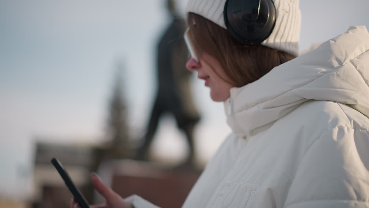 Youth grooving and tapping phone while wearing tinted goggles and headphones beside statue in snowy park during golden hour light, moving to rhythm in casual winter style against urban backdrop