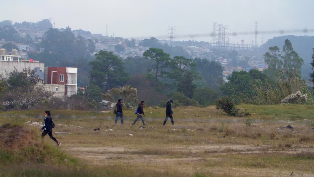 Street view with eerie atmosphere through barbed wire fence, people crossing field, favelas Zone 18 Guatemala City, Guatemala
