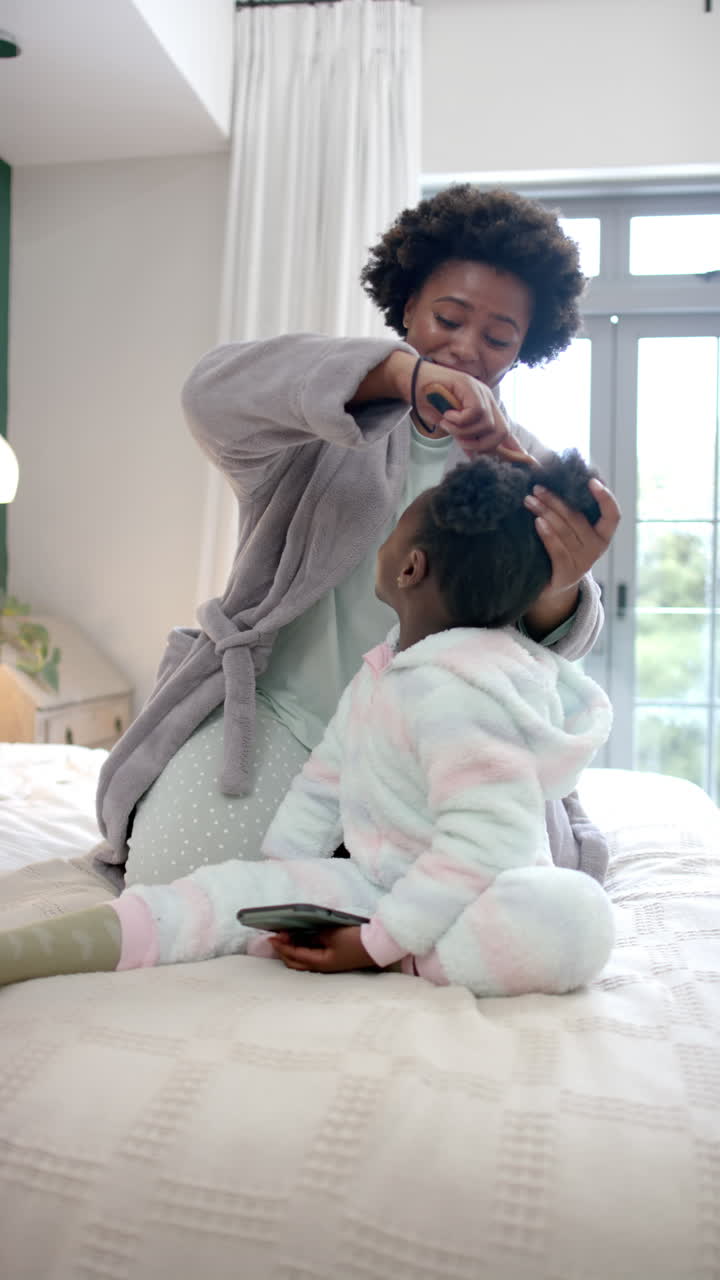 Vertical video of african american mother combing hair of daughter in bedroom, slow motion