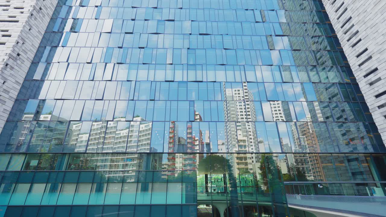 A low-angle shot of a towering modern glass facade, clearly reflecting the surrounding city buildings and the blue sky