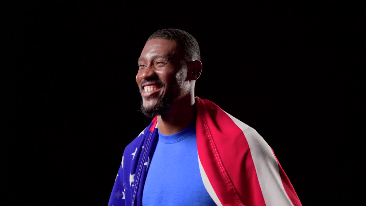 Uomo afroamericano adulto che celebra una vittoria, avvolto nella bandiera americana e con in mano un pallone da calcio, in studio.