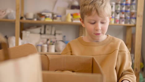 bambino biondo che guarda una bottiglia di plastica e la mette in una scatola di cartone su un tavolo in un laboratorio artigianale