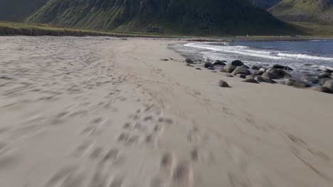 Aerial-shot-flying-close-to-the-sand,-revealing-waves-crushing-in-on-the-beach-and-a-mountain-in-the-background