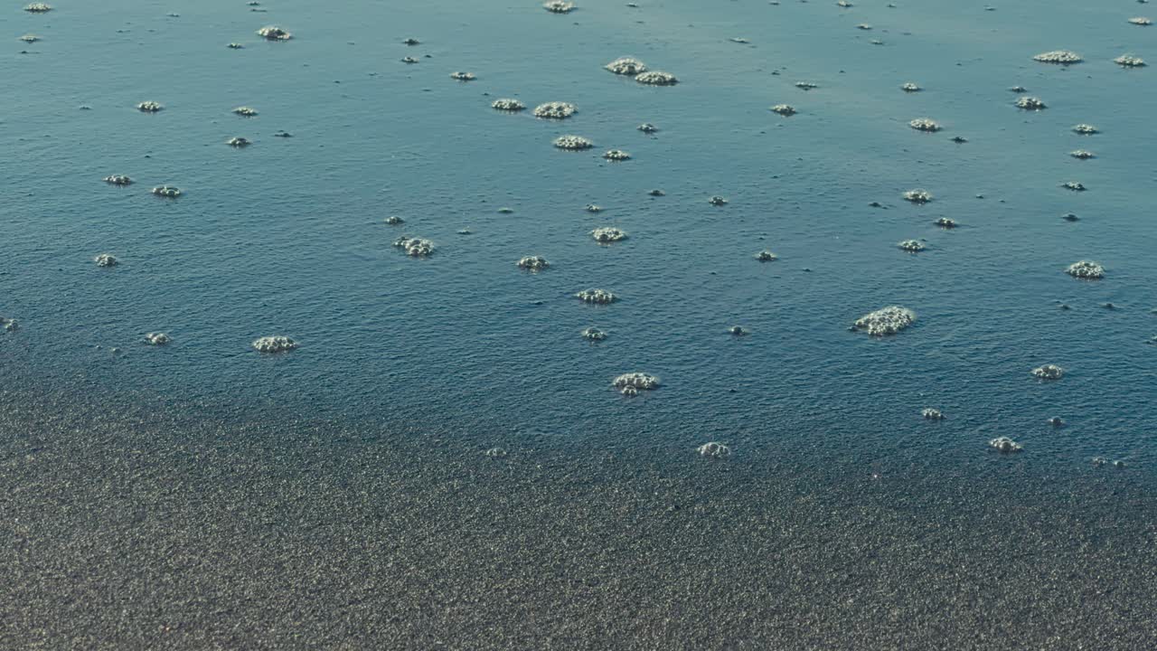 Bolle spumose dell'oceano formano motivi astratti su una spiaggia di sabbia nera mentre un'onda delicata entra nell'inquadratura, creando una scena naturalistica costiera cinematografica e artistica.