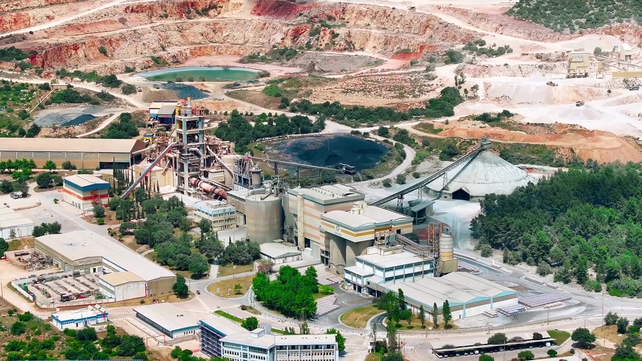 Aerial view of a cement production facility. Limestone quarry and aggregate processing operations are active. Heavy machinery including loaders and trucks transport materials.