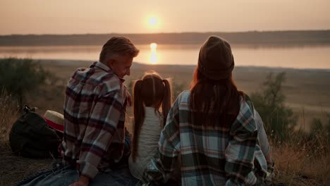 Rear-view-of-a-happy-married-couple-along-with-their-little-daughter-sitting-on-a-mat-during-their-picnic-and-enjoying-the-sunset-view-near-a-pond-in-the-summer-outside-the-city-in-the-evening