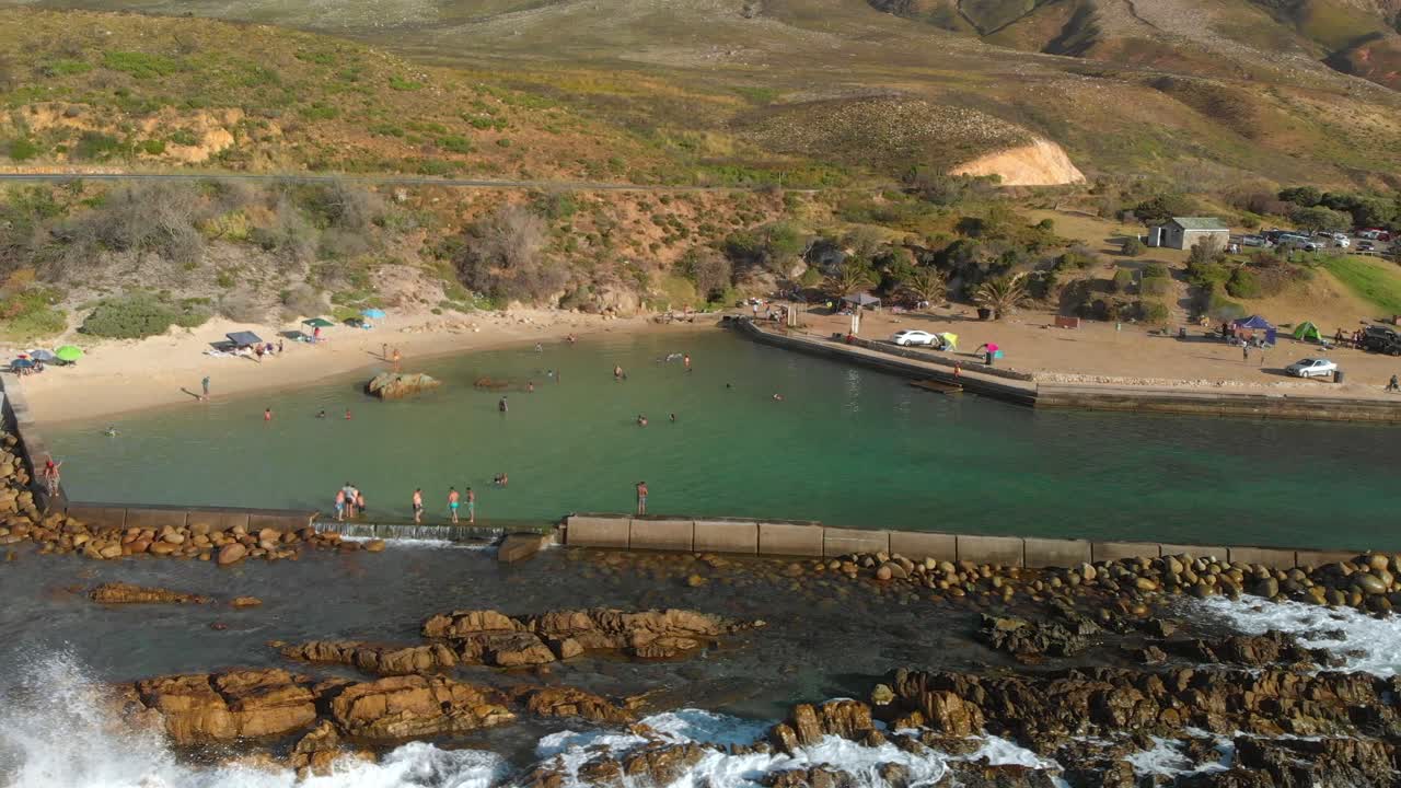 Drone view of ocean rock pool on rocky shoreline with people swimming with mountains in background