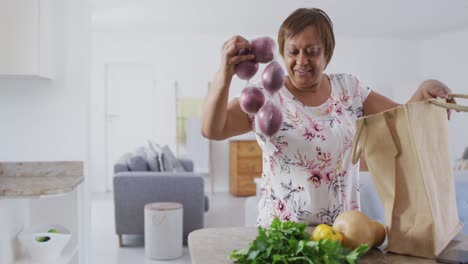 Feliz-Mujer-Mayor-Afroamericana-En-La-Cocina-Desempacando-Comida-De-La-Bolsa-De-Compras,-Sonriendo