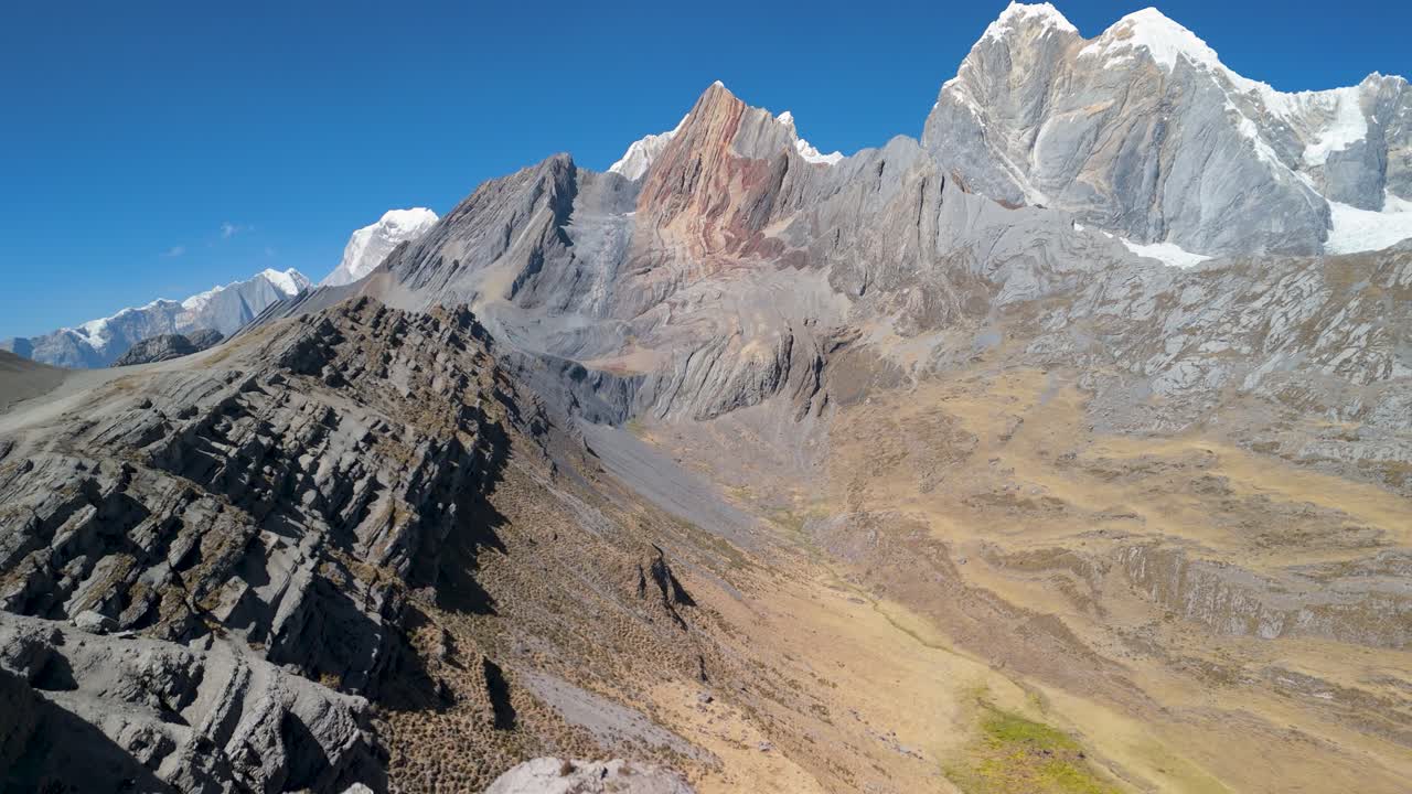 An epic aerial drone shot flies forward over the colorful, mineral-rich mountains of the remote Huayhuash trek, revealing the majestic snow-capped peaks of the Andes in Peru