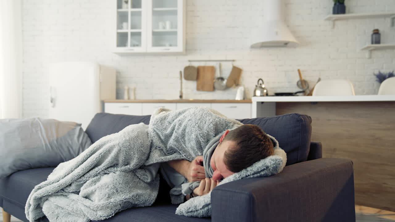 Young man trembling from fever. Sick man lying on couch at home and covering with warm blanket over the head.