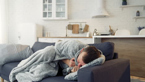 young man trembling from fever. sick man lying on couch at home and covering with warm blanket over the head.