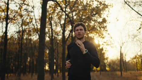 a confident man with curly hair and a beard in a black sports uniform runs along a forest path in an autumn forest with fallen brown leaves in the evening