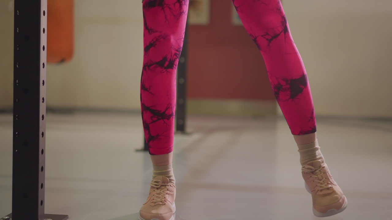 Calentamiento con banda de tobillo en estudio de gimnasio, primer plano de las piernas de atleta femenina con leggings rosados y zapatillas, dando pasos laterales con banda de resistencia alrededor de los tobillos, ritmo concentrado, jaula de sentadillas y entrenador.