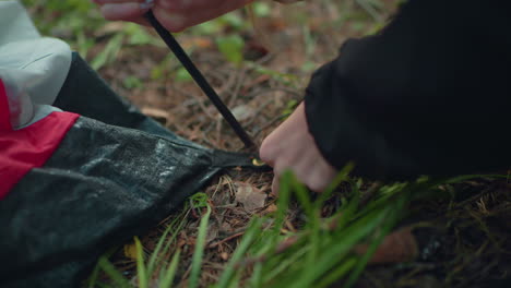 Close up of woman on forest floor picking up tent hook, forcing flexible pole through fabric hole and clipping gear into place, scene shows detailed motion of hands working on tent setup