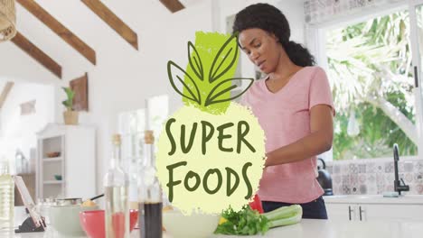Animation-Von-Blättern-Und-Superfood-Text-über-Einer-Afroamerikanerin,-Die-In-Der-Küche-Gemüse-Schneidet