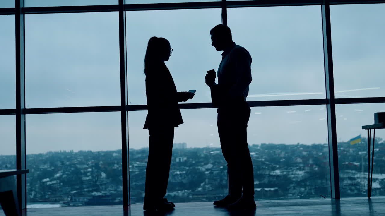 Dark silhouettes of male and female stand at the panoramic window. Conversation of colleagues in office. Low angle view.